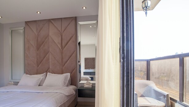 A modern bedroom with a large bed, a nightstand, a chair, and a view of the outdoors. - Darent Royal Threebedroom Al Malqa (Riyadh)