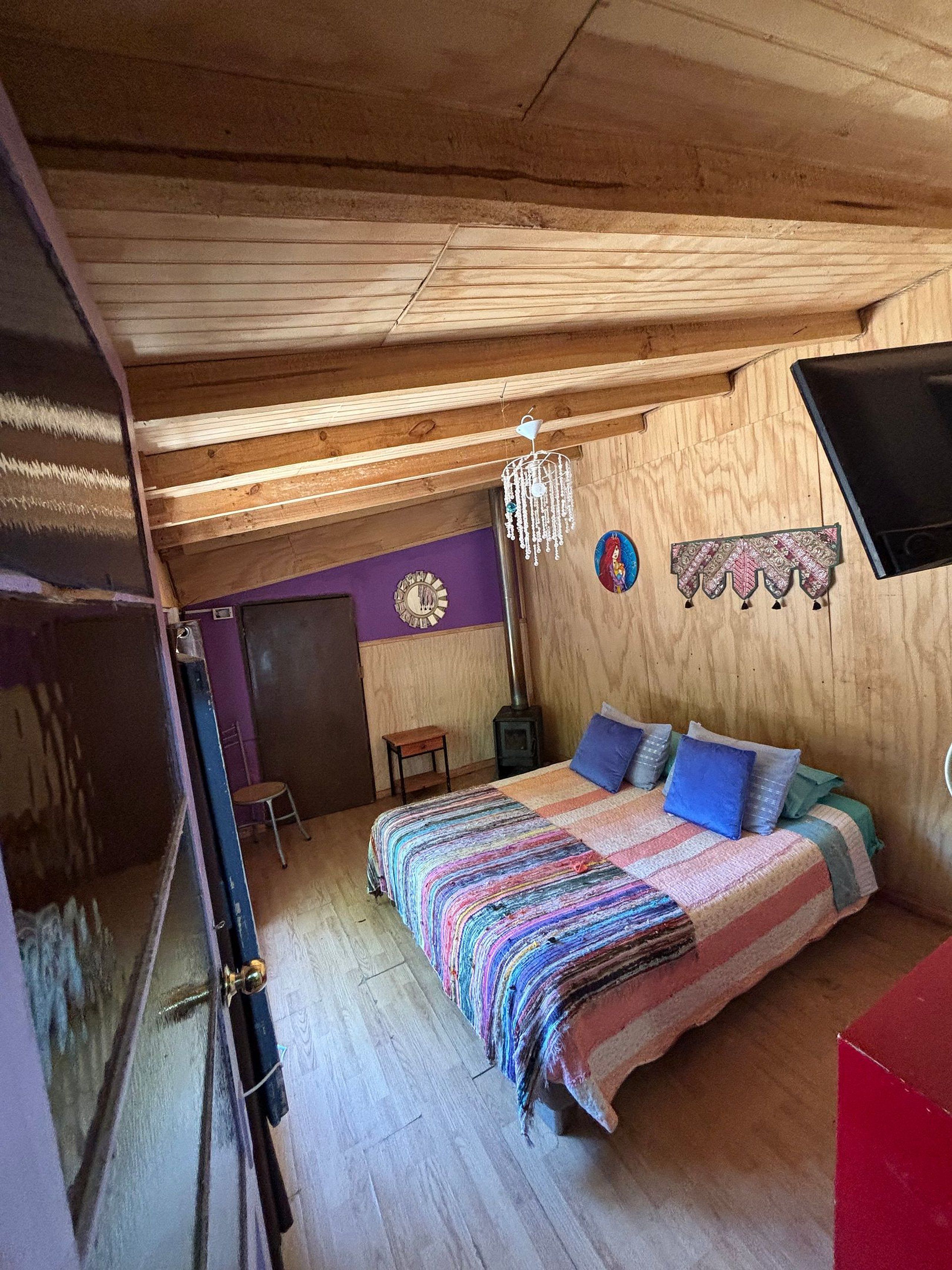 A cozy bedroom with a wooden ceiling, a bed with a colorful blanket, a small table, a wall-mounted TV, and a decorative wall hanging.