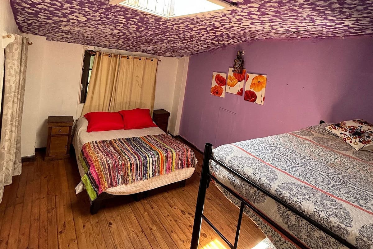 Ein Zimmer mit schrägem Dach, einem Oberlicht, einem Bett mit bunter Decke, einem Etagenbett und einem kleinen Nachttisch aus Holz.