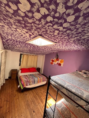Ein Zimmer mit schrägem Dach, einem Oberlicht, einem Bett mit bunter Decke, einem Etagenbett und einem kleinen Nachttisch aus Holz.