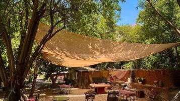 A hammock under a shade sail, outdoor furniture, and a rustic building in a garden setting.