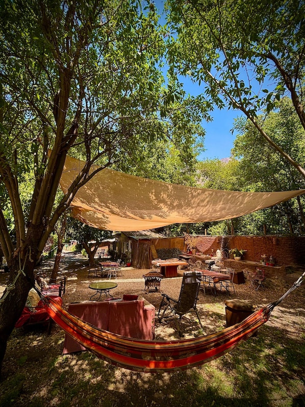 A hammock under a shade sail, outdoor furniture, and a rustic building in a garden setting. - Oceana Suites San San José de Maipo II (San Alfonso)