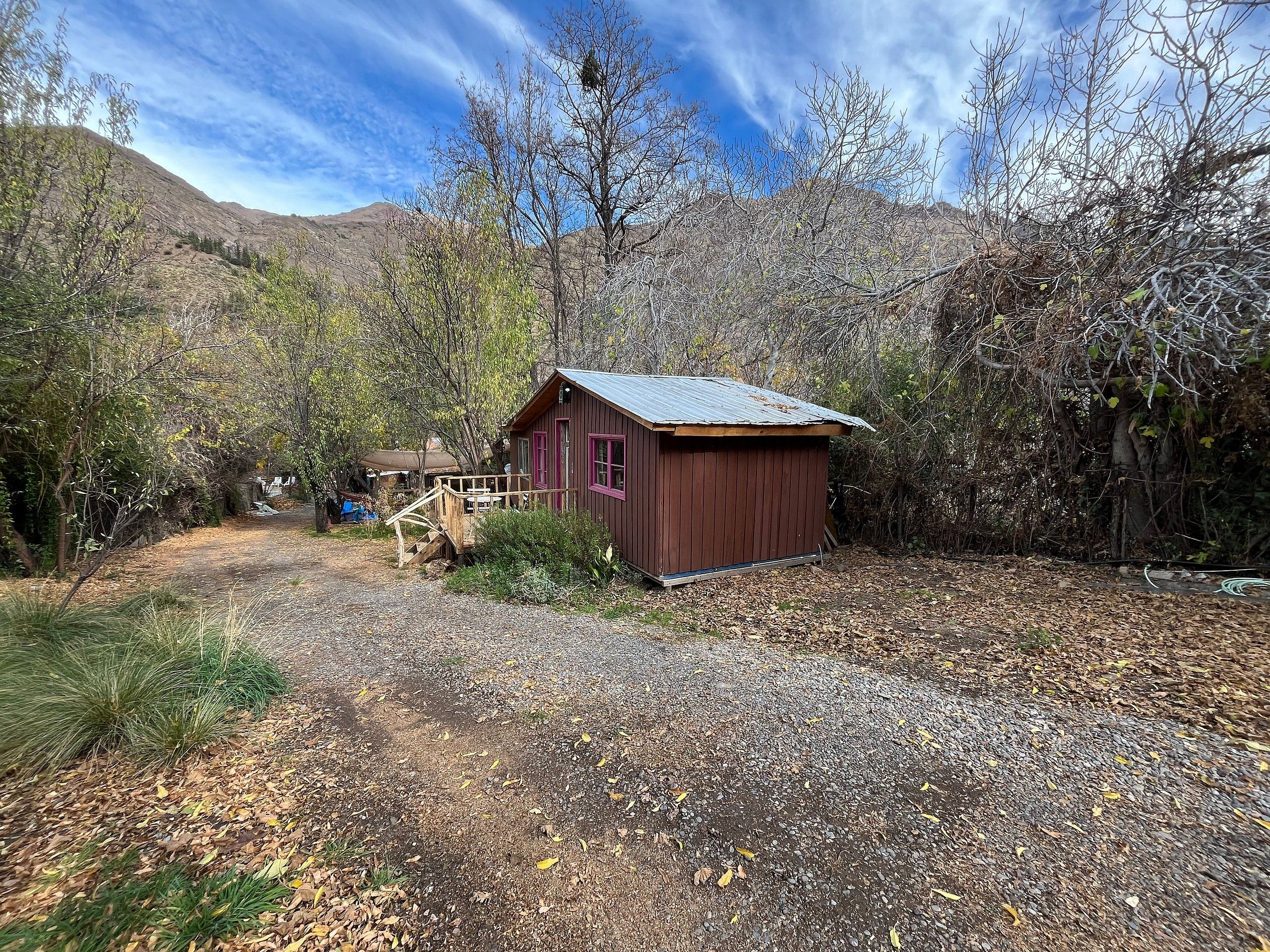 A rustic cabin with a wooden deck and a small porch, surrounded by trees and a gravel path.