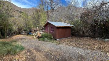 A rustic cabin with a wooden deck and a small porch, surrounded by trees and a gravel path.