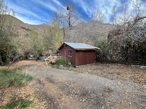 A rustic cabin with a wooden deck and a small porch, surrounded by trees and a gravel path. - Oceana Suites San San José de Maipo II (San Alfonso)