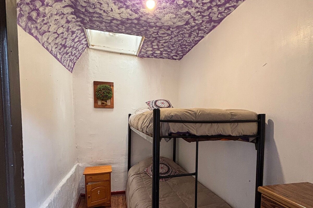 Habitación con literas, techo con estampado floral, una ventana pequeña y una mesita de noche de madera.