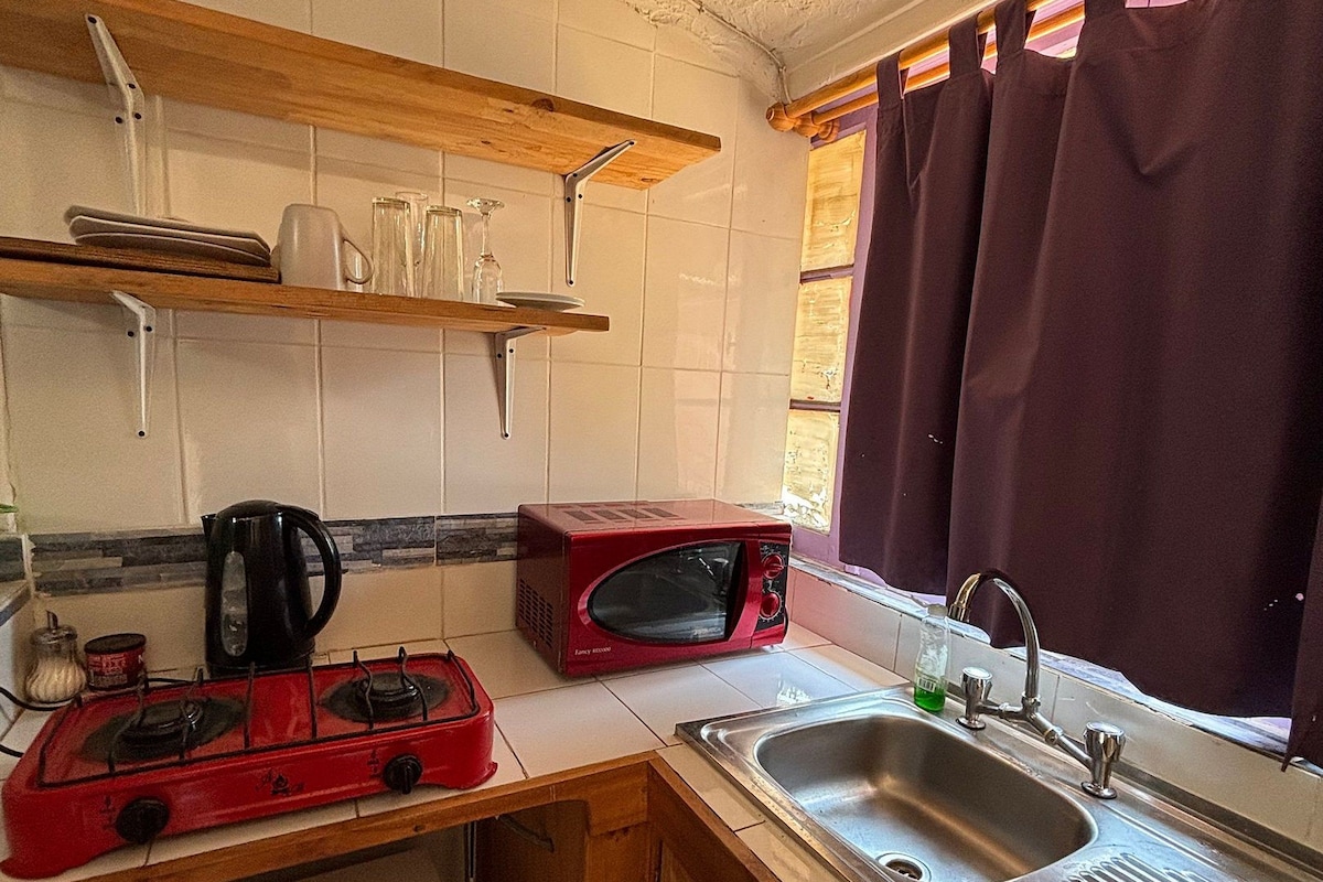 Una cocina compacta con estufa a gas, un microondas rojo, un lavamanos y una ventana con cortinas moradas.