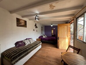 A room with a wooden floor, a sofa with patterned cushions, a wooden table, and a window with a view of trees. - Oceana Suites San San José de Maipo II (San Alfonso)