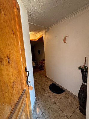 Un pasillo con puerta de madera, suelo de baldosas, felpudo negro, decoración de luna creciente en la pared y una cesta de mimbre con flores.