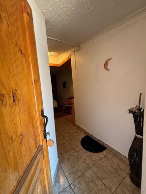 Ein Flur mit einer Holztür, einem Fliesenboden, einer schwarzen Matte, einer Wanddekoration im Halbmond-Design und einem Korbwägelchen mit Blumen.