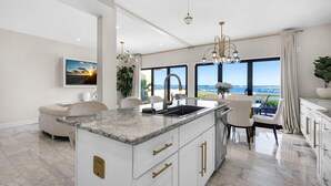 Een moderne keuken met een groot eiland, witte kastjes en gouden handgrepen. Er is een zithoek met witte stoelen en uitzicht op zee door grote ramen.