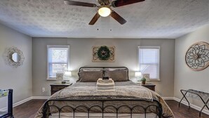 A bedroom with a bed, a ceiling fan, two bedside tables, a nightstand, a wall-mounted decorative item, and a window with blinds.