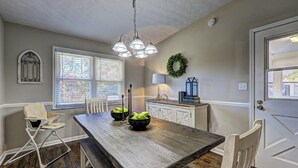 A dining area with a wooden table, chairs, a chandelier, a window with blinds, a door, and a wall-mounted wreath.