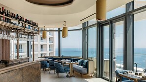 Un bar moderno con botellas de vino y copas, un espacio de estar con sofás y mesas, y amplias ventanas con vistas al océano.