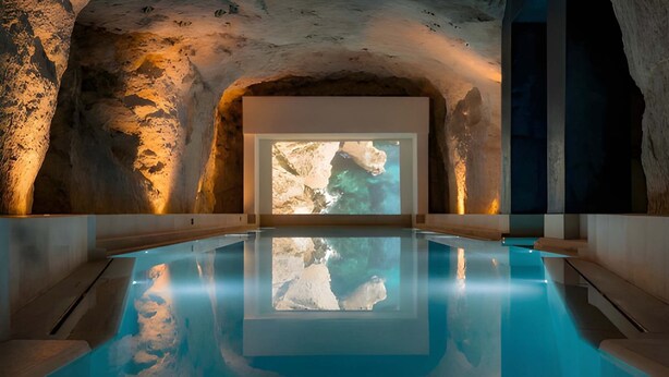 Una piscina interior con techo que imita una cueva rocosa y una gran pantalla mostrando una escena costera.
