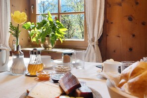 A breakfast table with various food items, a vase with a yellow flower, and a window with a view of trees.