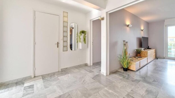 En modern inredning med en hall som leder till ett vardagsrum, med bland annat en vit dörr, en spegel, en växt, en tv och en träkonsol.
