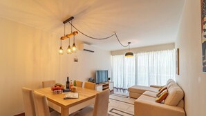 A modern living room with a dining area, featuring a wooden table, beige sofa, and pendant lighting. - Home Azores - Loreto Apartment (Ponta Delgada)