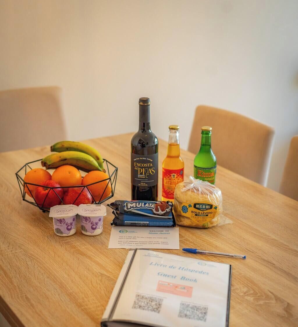 Uma mesa com uma garrafa de vinho Encosta de Pias, uma garrafa de sumo de laranja, uma garrafa de água verde, pão embrulhado, uma taça de fruta e um livro de visitas.