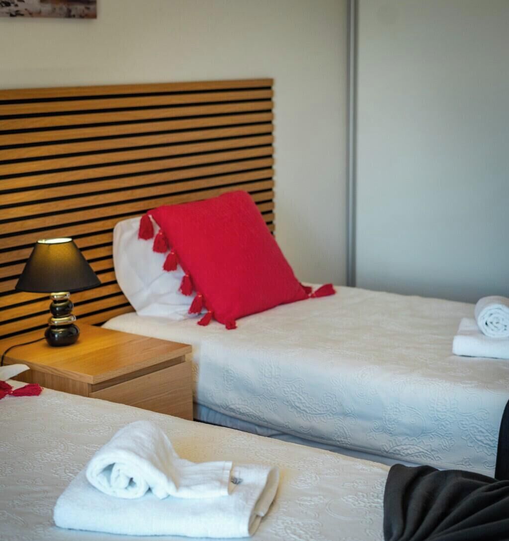 Hotellihuone, jossa on kaksi sänkyä, yöpöytä lampun kanssa, punainen tyyny ja rullalle taitetut pyyhkeet.
