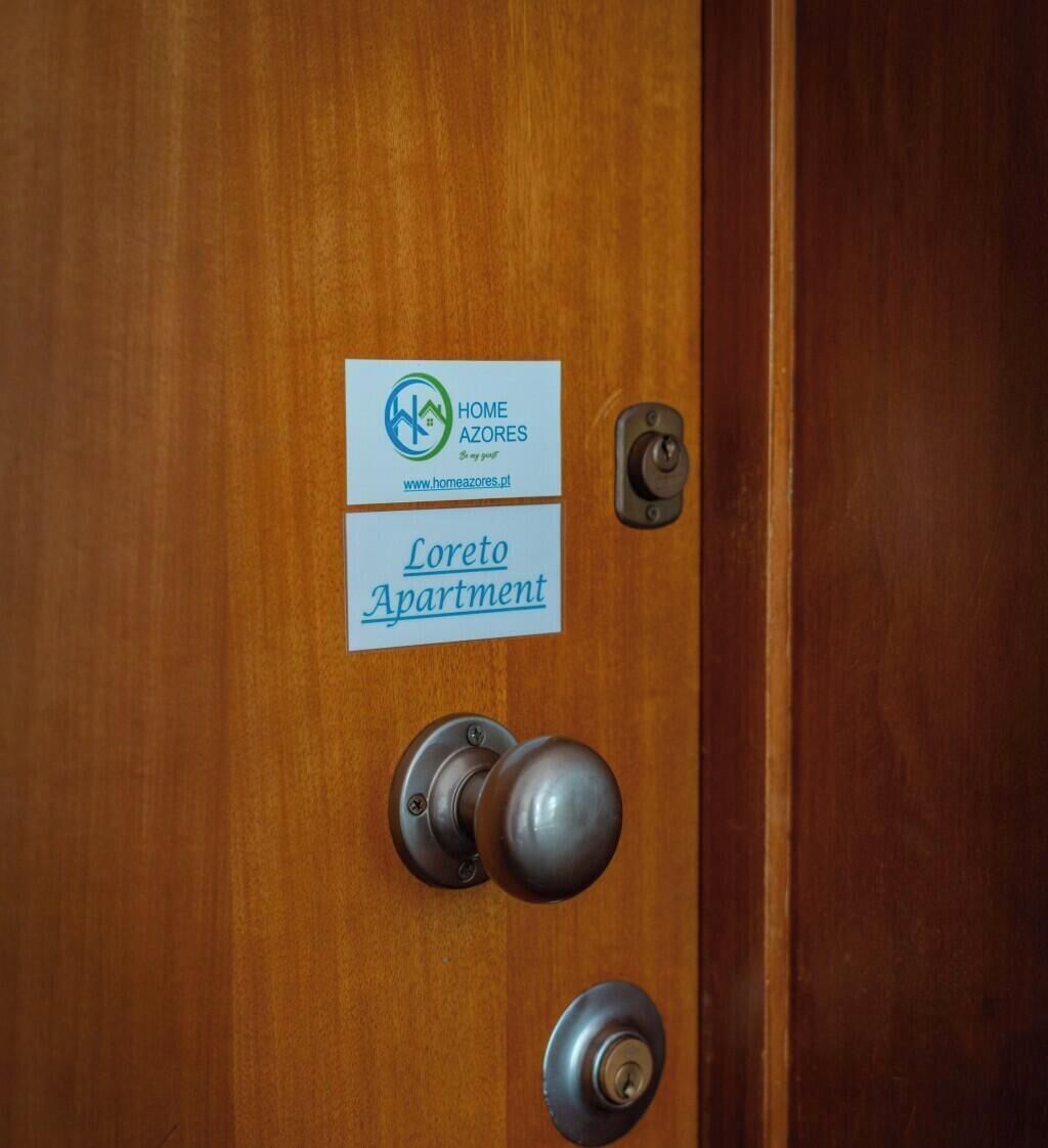 Uma porta de madeira com maçaneta de metal, uma fechadura e duas placas: uma com 'Home Azores' e outra com 'Loreto Apartment'.
