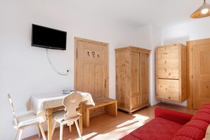 Quarto com televisão na parede, armários de madeira, uma mesa pequena com cadeiras e um sofá vermelho.