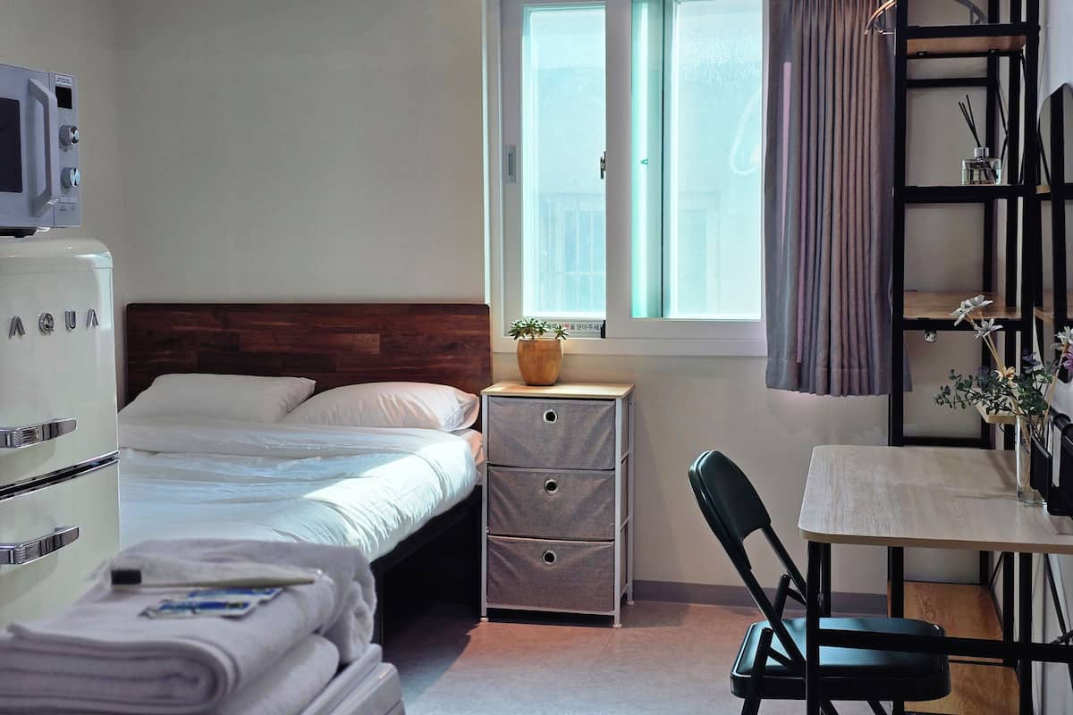 A bedroom with a bed, a desk, a chair, a nightstand, a dresser, a window with curtains, and a potted plant.