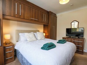 A bedroom with a bed, a television, wooden cabinets, a nightstand with a lamp, and a mirror. - The Nook (Whitby)
