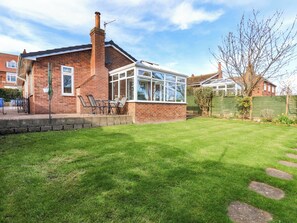 A brick house with a conservatory, a lawn, and a patio with chairs. - The Nook (Whitby)