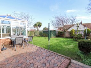 A patio with a table and chairs, a conservatory, a well-maintained lawn, and a house with a tiled roof. - The Nook (Whitby)