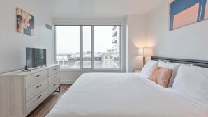 A modern hotel room with a large bed, a TV on a dresser, a window with a city view, and a painting on the wall. - Modern Apt Heart of Seaport (Boston, MA)