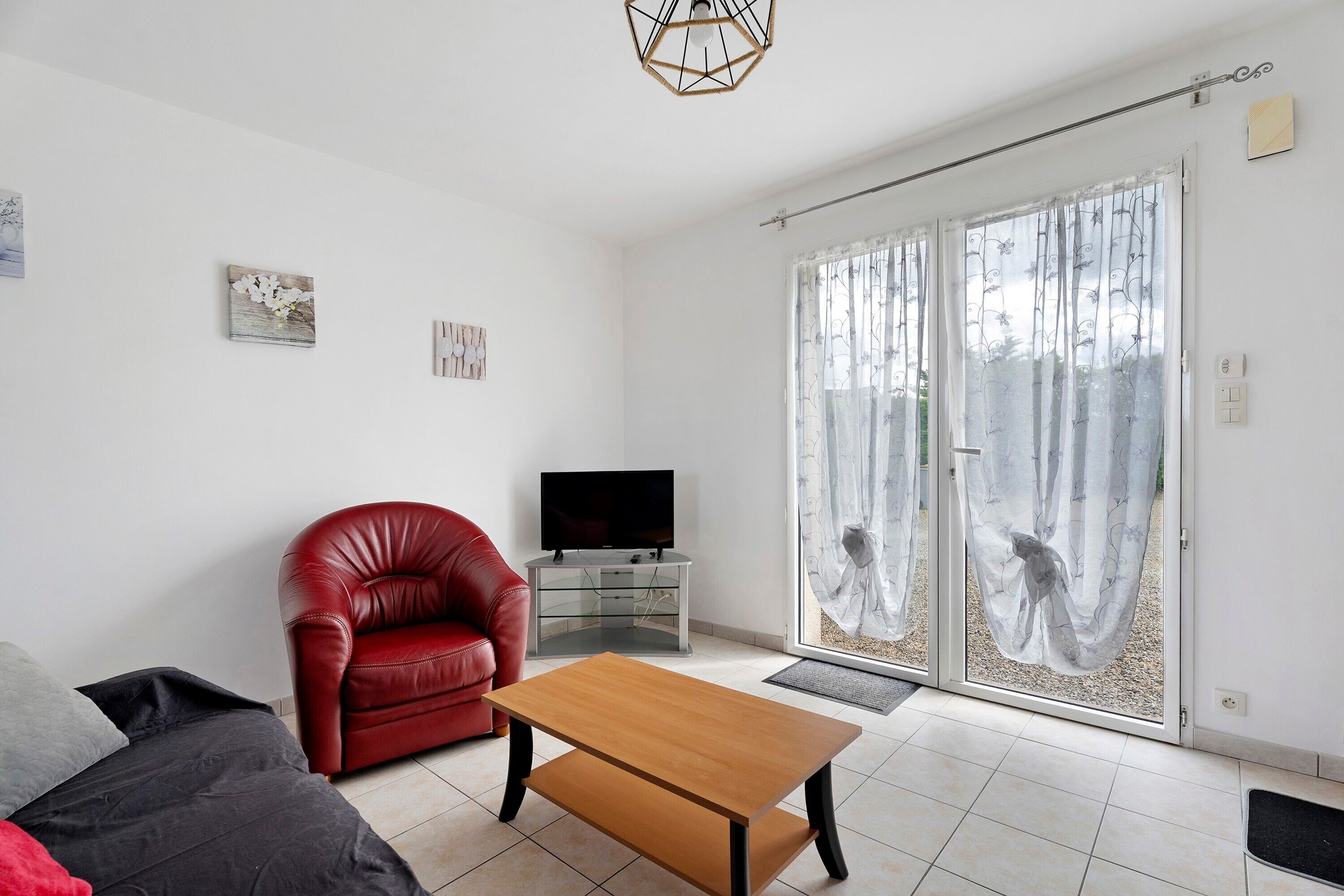 A living room with a red leather chair, a wooden coffee table, a TV on a glass cabinet, and a sliding glass door with curtains.