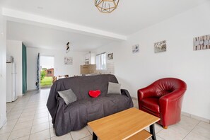 Una sala de estar con sofá, una silla roja, una mesa de madera y una cocina al fondo.