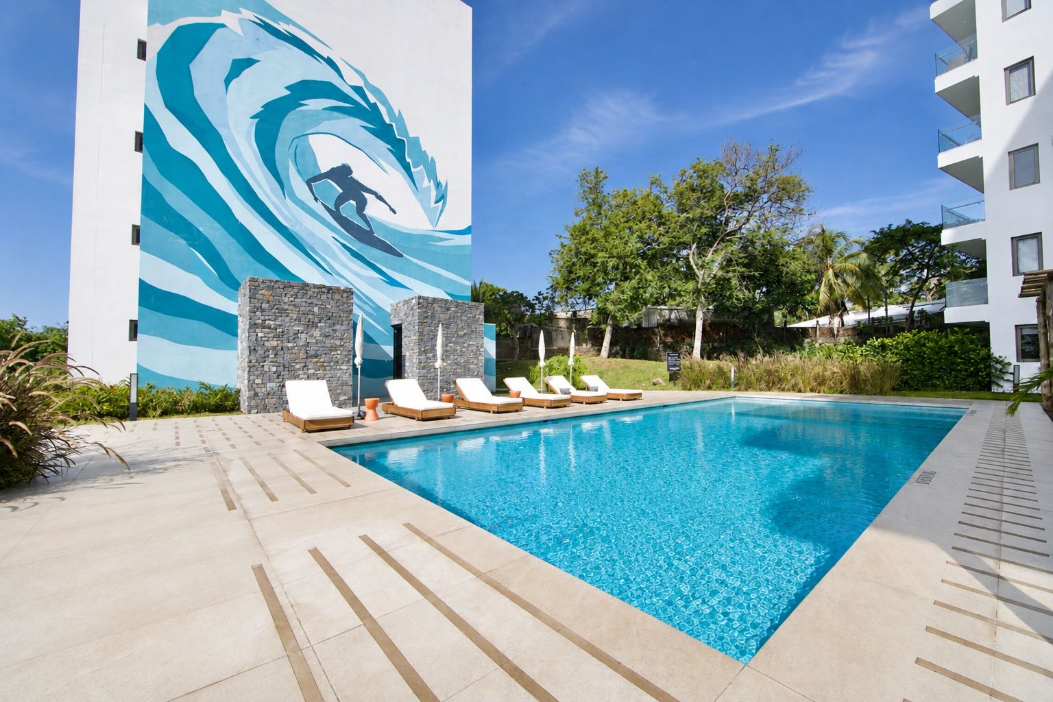 A swimming pool with lounge chairs, a stone wall, and a building with a wave mural.