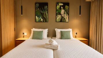 A double bed with white bedding and green pillows, two wall-mounted paintings of foliage, wooden nightstands with lamps, and a wooden wardrobe.