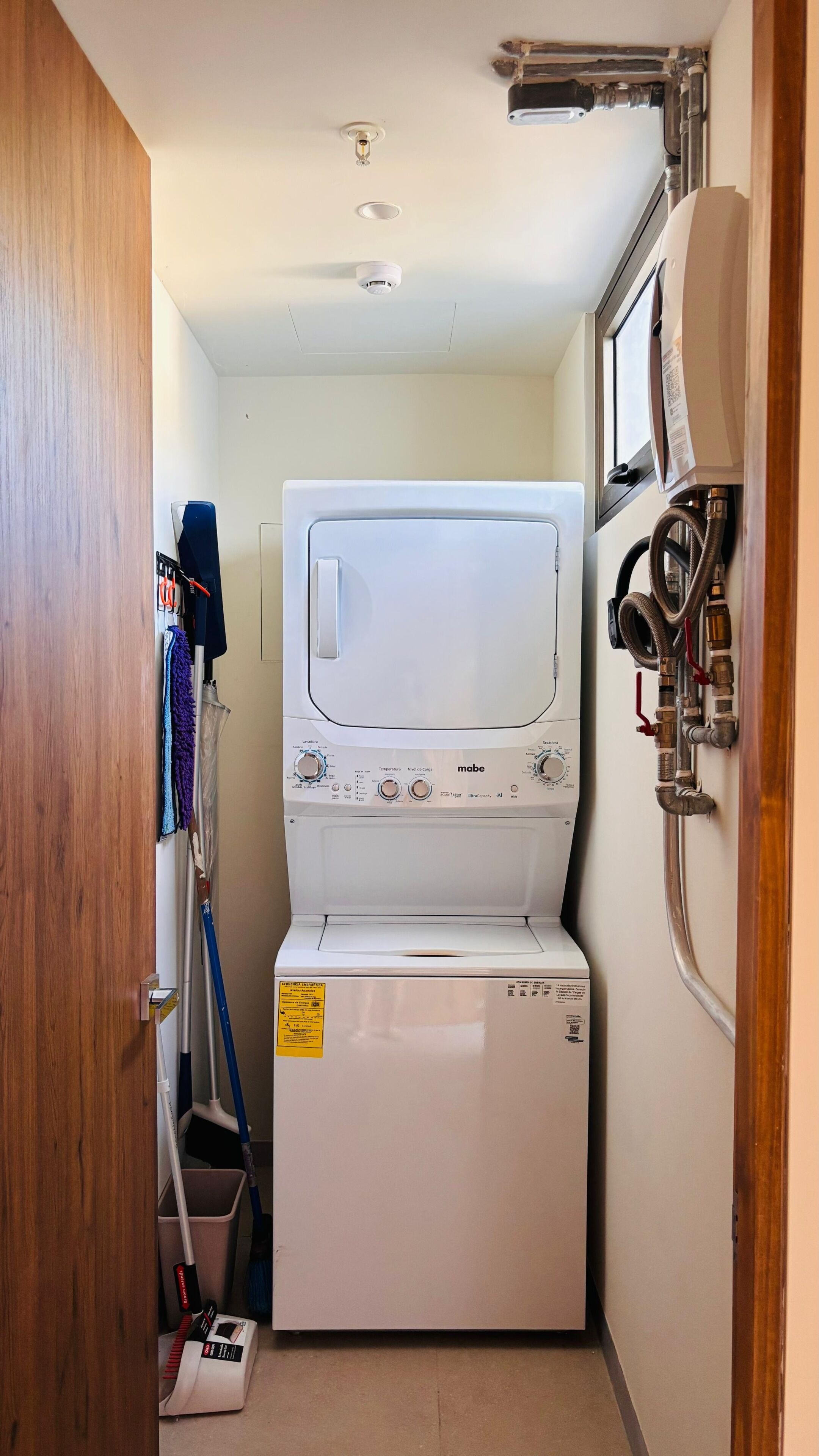 A compact laundry room with a stacked washer and dryer, a mop and broom, and visible plumbing.
