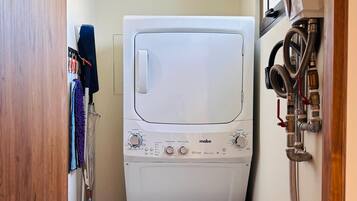 A compact laundry room with a stacked washer and dryer, a mop and broom, and visible plumbing.