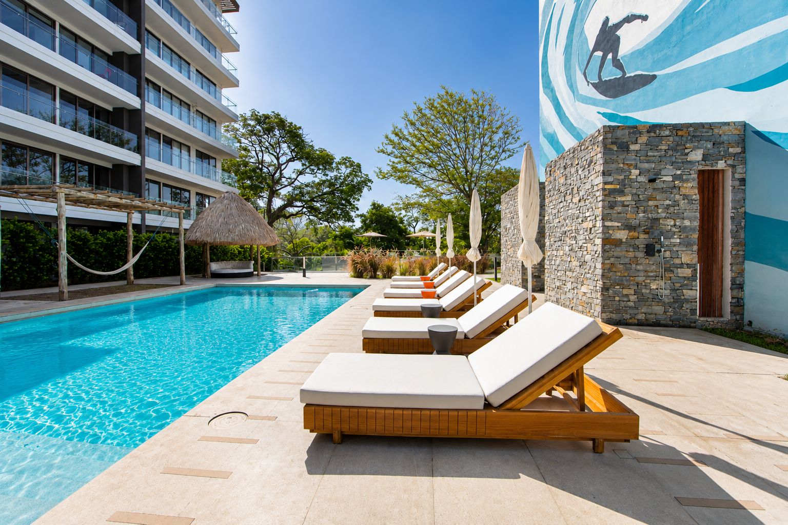 A swimming pool with lounge chairs and umbrellas, a stone wall, and a mural of a surfer.