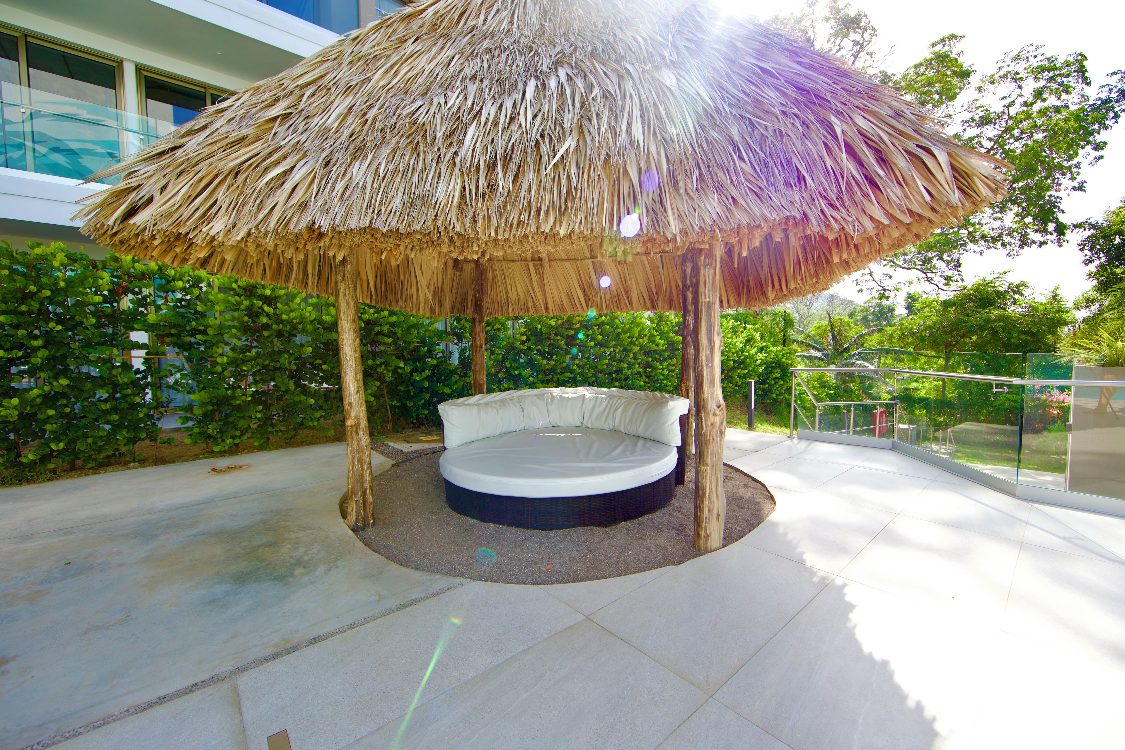 A thatched-roof gazebo with a circular seating area, surrounded by greenery and a building in the background.