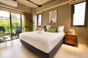 A modern bedroom with a large bed, a bedside table, a chair, and a view of greenery outside. - El Zonte Garden 101,  Private Garden, Steps to Pool and Surf. (El Zonte)