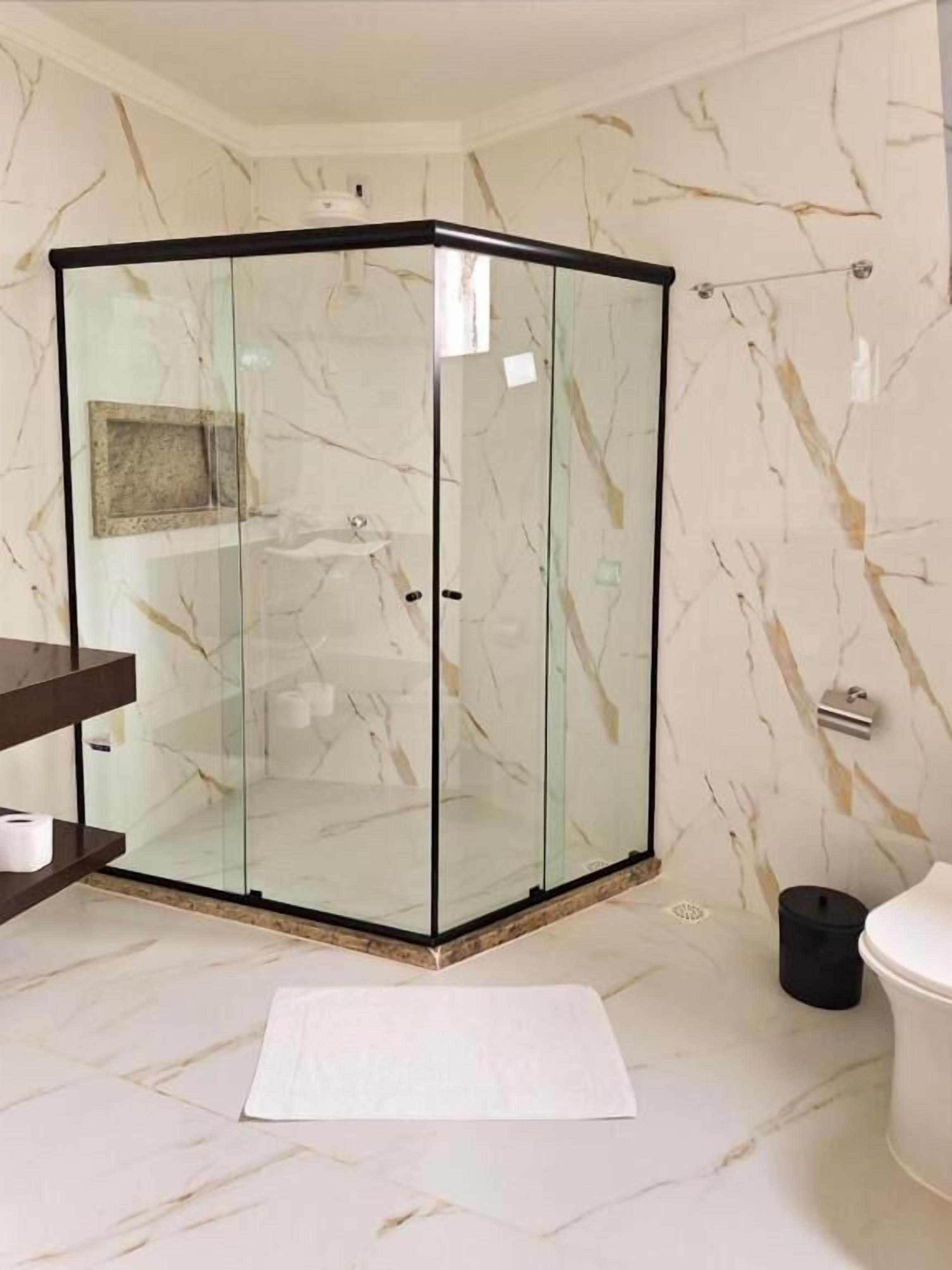 Una ducha en esquina con puertas de cristal, pared con diseño de mármol, suelo blanco y un cesto de basura negro.