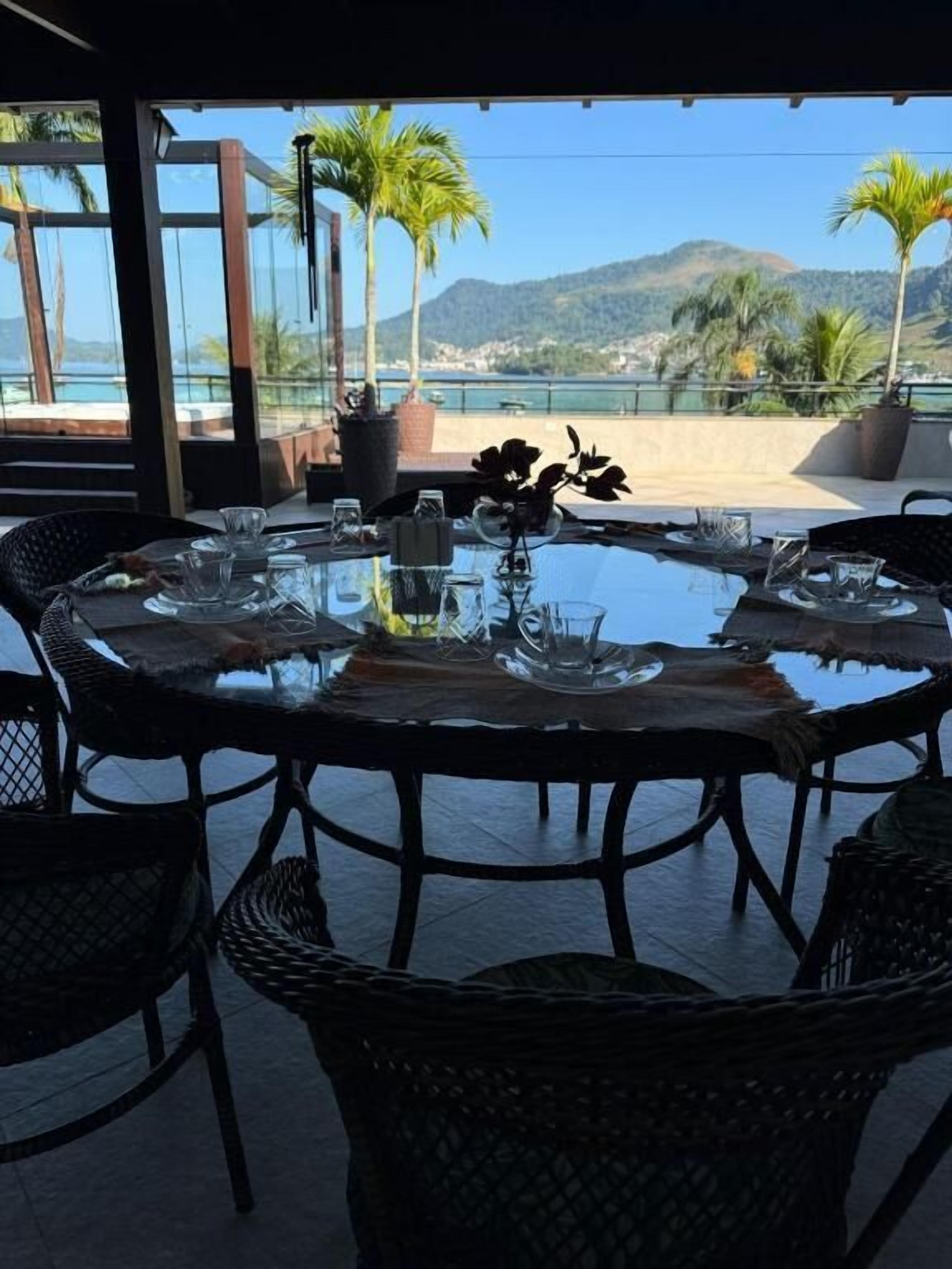 A round table with chairs, a glass top, and a view of the sea and mountains.