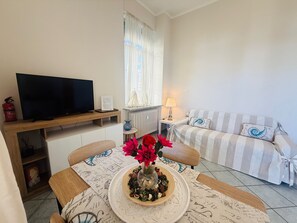 Ein Wohnzimmer mit Fernseher, einer Couch, einem Tisch mit Blumenvase und einem Fenster mit Vorhängen.