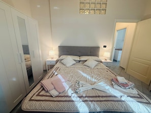 Ein ordentlich bezogenes Bett mit einem gemusterten Überwurf, zwei Nachttische mit Lampen, ein Spiegel und ein Kleiderschrank.