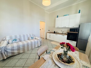 Ein modernes Wohnzimmer mit einer Couch, einem Esstisch mit Blumenvase und einer Küchenzeile mit Schränken und Geräten.