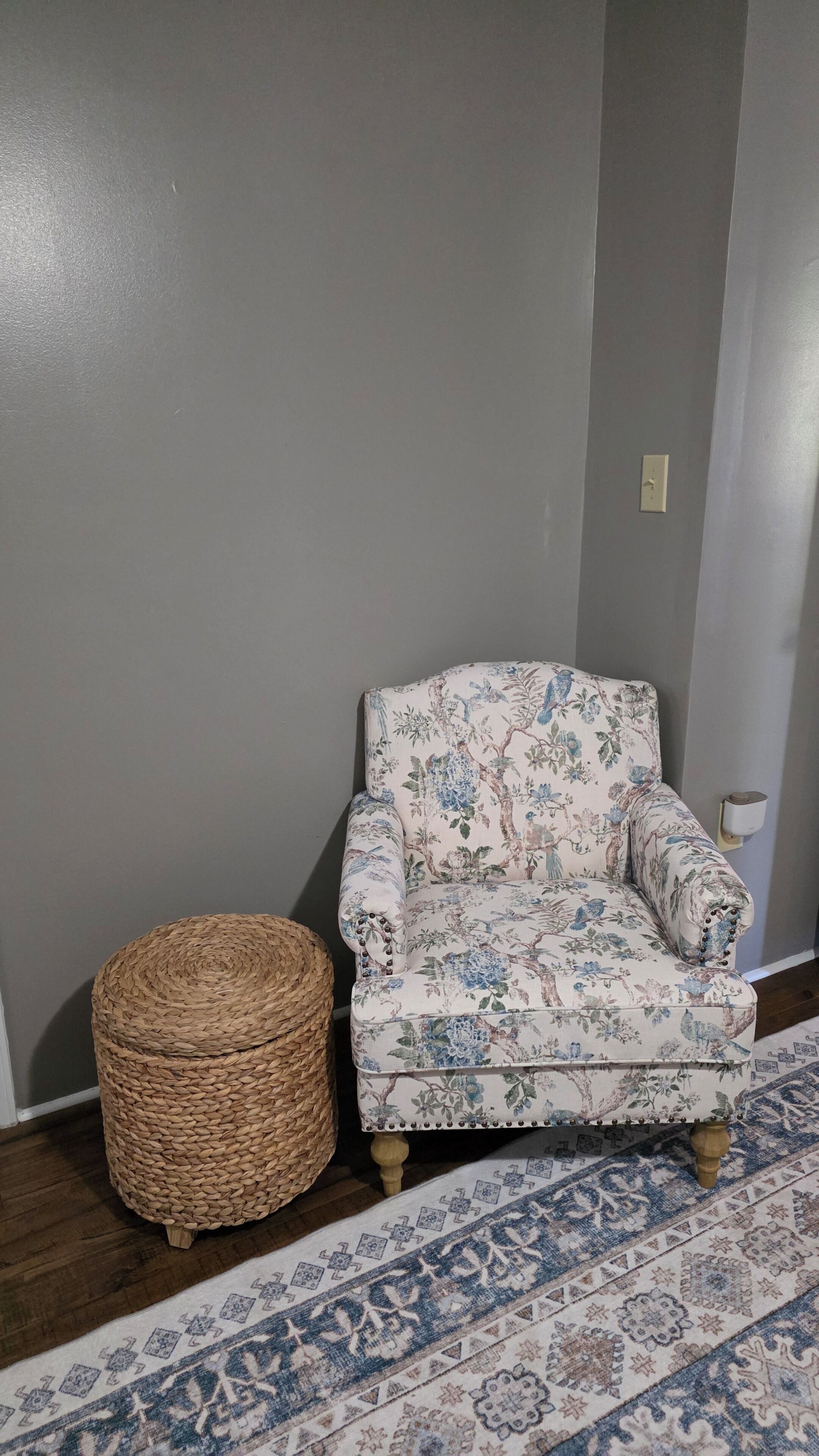 A room with a grey wall, a floral armchair, a woven ottoman, and a patterned rug.