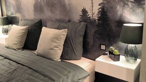 Un dormitorio con cama, almohadas, una mesita de noche, una lámpara y un mural en la pared con un bosque.