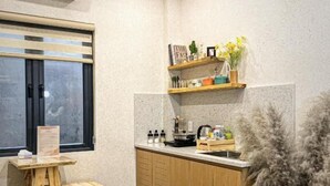 Una cocina compacta con armarios de madera, una estantería empotrada con objetos decorativos, una pequeña mesa de madera y una ventana con persianas.