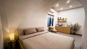 Un dormitorio con una cama grande, una mesita de noche con una lámpara, un taburete pequeño y vistas a una zona de cocina con armarios y una ventana.