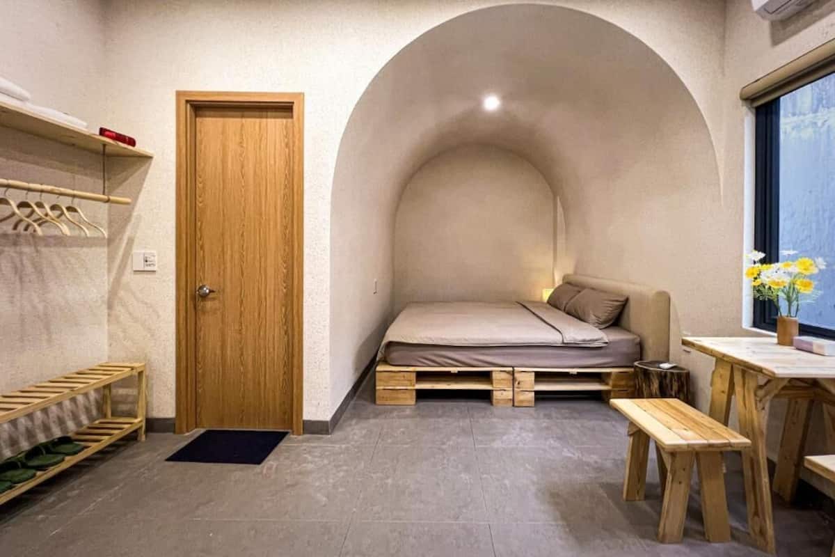Una habitación pequeña con cama entrepiso, puerta de madera, un banco, una mesita y ventana con vistas.
